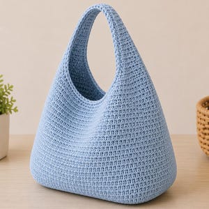 Op de afbeelding: Een lichtblauwe gehaakte handtas met een driehoekig silhouet en een enkel geïntegreerd handvat. De tas heeft een dicht, uniform steekpatroon over de gehele buitenkant, wat zorgt voor een textuur met een brede basis en een taps toelopende opening.