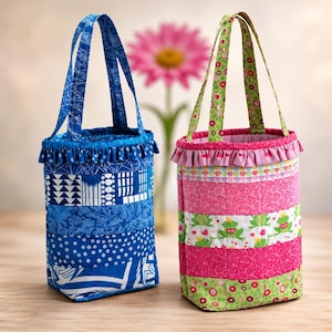 May include: Two cylindrical fabric bags with ruffled tops and dual handles. The left bag features various blue patterns including geometric shapes and dots. The right bag displays pink heart patterns, green frog illustrations, and floral accents.