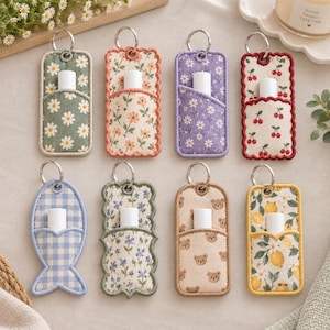 May include: Eight rectangular fabric lip balm holders with metal key rings, featuring various patterns including daisies, cherries, lemons, bears, and blue gingham. Each holder has a small pocket containing a white cylindrical lip balm tube.