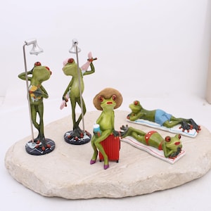 May include: A set of five green frog figurines in various poses. Two frogs stand under silver shower fixtures. One frog sits on a red suitcase wearing a tan hat and holding a blue cup. Two frogs lie on white mats, one wearing blue shorts and one in red polka-dot bottoms.