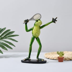 Op de afbeelding: Een groen kikkerbeeldje op een zwarte voet, dat een zilveren tennisracket in één hand houdt met de andere arm uitgestrekt. De kikker heeft een lichtgroene buik met donkergroene vlekken en donkergroene ledematen in een dynamische houding.