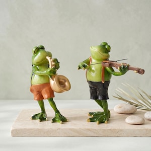 Puede incluir: Dos figuras de ranas verdes de pie. La rana de la izquierda lleva pantalones cortos naranjas y toca un saxofón dorado. La rana de la derecha lleva pantalones cortos negros con tirantes marrones y toca un violín marrón con un arco negro.