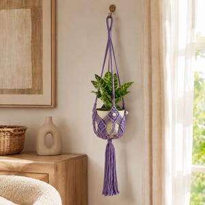 May include: A lavender macrame plant hanger featuring a knotted cord design with small wooden beads, holding a white ceramic pot containing a green leafy plant. The hanger includes a long, thick tassel at the bottom and a loop for hanging.