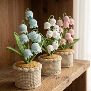 Könnte beinhalten: Drei gehäkelte Maiglöckchen-Pflanzen in beigen Töpfen mit gelben und gewellten Verzierungen. Die Blüten sind hellblau, weiß und rosa, mit grünen Blättern und Stielen. Die Pflanzen werden auf einem Holzregal ausgestellt.