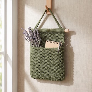May include: A sage green macrame wall hanging pocket made of thick cord, suspended from a light wooden dowel and a matching cord loop. The pocket contains a bundle of dried purple lavender stems, a small spiral-bound notebook, and a silver pen.