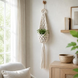 May include: A cream-coloured macrame plant hanger featuring an intricate knotted net design, a circular wooden ring at the top for suspension, and a long, thick tassel at the bottom. A small green potted plant is nestled inside the woven structure.