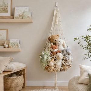 May include: A cream-colored macrame toy hammock hanging from a wall hook, filled with a collection of plush stuffed animals including a brown bear, a white rabbit, a fox, a green dinosaur, a giraffe, a sheep, an elephant, and an owl.