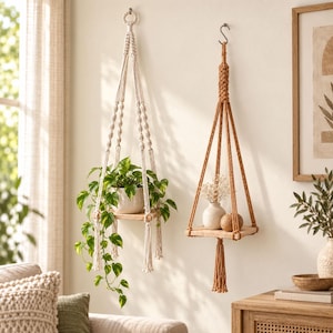 Peut inclure: Deux suspensions pour plantes en macramé fixées au mur. La suspension de gauche est blanc cassé avec une étagère en bois carrée portant une plante pothos verte. Celle de droite est terracotta avec une étagère en bois portant un vase blanc.