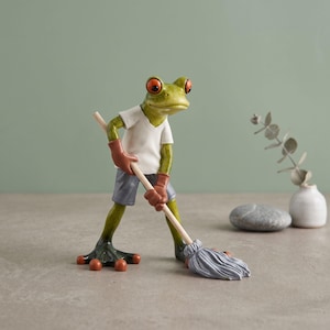 Könnte beinhalten: Eine grüne Froschfigur mit großen orangefarbenen Augen, bekleidet mit einem weißen V-Ausschnitt-T-Shirt, grauen Shorts und braunen Handschuhen. Der Frosch hält einen hölzernen Wischmopp mit grauem Kopf und steht in einer Putzpose.