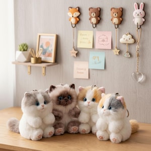 May include: Four plush cat keychains in white, gray, brown, and orange. Each cat has big blue eyes and a fluffy tail. Decorative hooks shaped like a fox, bears, and a bunny hang above the cats. A small shelf with a plant and picture frame is also visible.