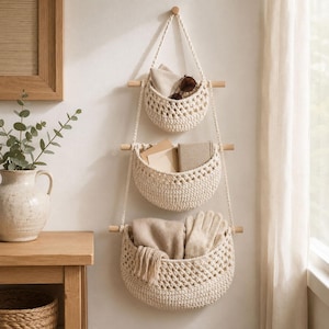 May include: A three-tiered wall-hanging storage unit featuring cream-coloured crocheted baskets suspended by ropes from three horizontal wooden dowels. The baskets contain various items including a pair of sunglasses, papers, and a pair of gloves.