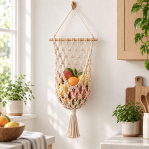 May include: A cream-coloured macrame fruit hammock featuring a diamond-patterned knotted cord design suspended from a wooden dowel. The hanging storage basket holds a variety of fresh fruits including oranges, apples, and lemons, finished with a tassel.