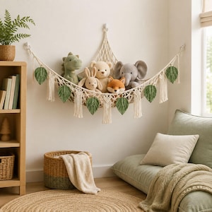 May include: A cream-colored macrame toy hammock suspended on a wall, featuring decorative green fabric leaves and fringe tassels. The hammock holds five plush stuffed animals, including a dinosaur, a rabbit, a bear, a fox, and an elephant.