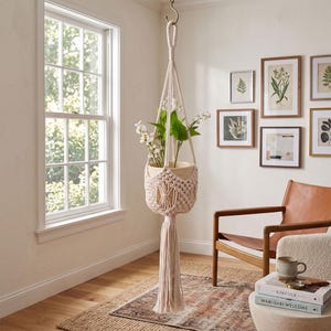 May include: A cream-coloured macrame plant hanger featuring intricate knotted patterns and a long, thick fringe tassel at the bottom. The hanger holds a light-toned wooden pot containing white artificial flowers with green leaves, suspended by a hook.