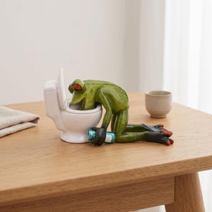 Peut inclure: Une figurine de grenouille verte aux yeux orange et aux doigts rouges, penchée sur une cuvette de toilettes en céramique blanche. La grenouille tient un petit objet cylindrique bleu et argenté dans une main, le corps en position agenouillée.