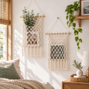 Op de afbeelding: Twee off-white macramé wandhangers met een geknoopt netontwerp opgehangen aan houten stokken. Elk stuk bevat een rechthoekig framevak met franjes aan de onderkant, waarin respectievelijk groen blad en een ingelijste foto zijn geplaatst.