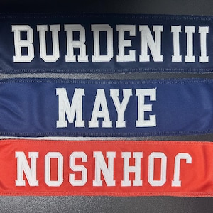 May include: Three rectangular patches in navy blue and orange, each with white block lettering. The top patch reads "BURDEN III", the middle patch says "MAYE", and the bottom patch says "NOSNHOJ". These patches are likely for sports or team apparel.