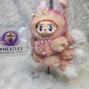 May include: A plush keychain figure with pink and peach faux fur, featuring a wide-eyed face, a toothy grin, and pointed ears. It is adorned with a beaded necklace with a pink butterfly charm, a sheer butterfly hair clip, and a metal keyring.
