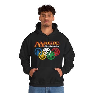 May include: Black hooded sweatshirt with the text "MAGIC The Gathering" in gold and red. Below the text are five colored rings with symbols. The sweatshirt has a front pocket and a drawstring hood.