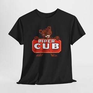 Puede incluir: Camiseta negra con un diseño vintage. El diseño muestra un oso pardo sosteniendo un letrero rojo con las palabras "PIPER CUB" en blanco. Debajo, se lee "PIPER AIRCRAFT CORP. VERO BEACH, FL U.S.A.".