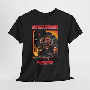 May include: Black t-shirt with a movie poster design. The poster features the text "SCHWARZENEGGER" in red at the top and "PREDATOR" at the bottom. The artwork shows Arnold Schwarzenegger and other characters from the film.