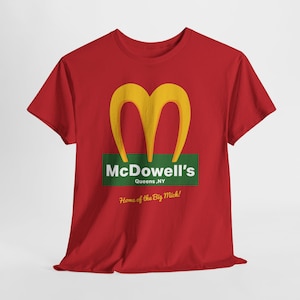 May include: Red t-shirt featuring a yellow double arch logo above the text "McDowell's Queens, NY" on a green rectangle. Below, the text reads "Home of the Big Mick!"