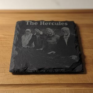 May include: A black slate coaster with the engraved text "The Hercules" and a group photo of four people. The coaster has a rough, natural edge and sits on a wooden surface.