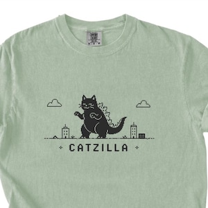 May include: A light sage green t-shirt featuring a black pixel art graphic of a cat resembling a monster standing among small buildings and clouds. Below the graphic, the text CATZILLA is printed in a bold, pixelated black font with small crosses.