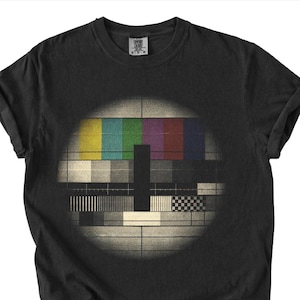 May include: Black t-shirt featuring a retro television test pattern design. The graphic includes colorful bars and grayscale blocks within a circular frame. The shirt has rolled-up sleeves and a crew neck.