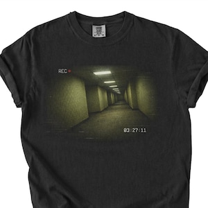 Peut inclure: T-shirt noir avec un graphisme représentant un long couloir faiblement éclairé avec des lumières au plafond. Le mot "REC" est en rouge en haut à gauche, et l'heure "03:27:11" est affichée en bas. Le t-shirt a des manches retroussées.