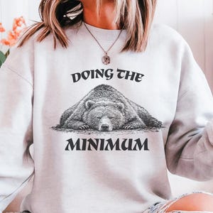 Puede incluir: Una sudadera gris claro con el texto "DOING THE MINIMUM" encima y debajo de una ilustración detallada de un oso durmiendo. El oso es en tonos grises y marrones.