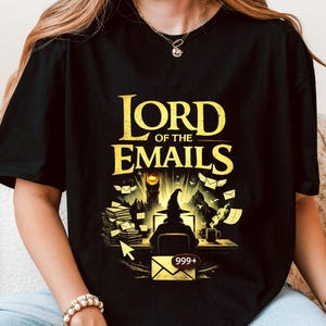 Peut inclure: T-shirt noir avec un motif doré qui dit "LORD OF THE EMAILS". Le motif représente un sorcier devant un ordinateur avec une pile d'e-mails, une tour et une tasse de café. L'icône de l'e-mail affiche "999+".