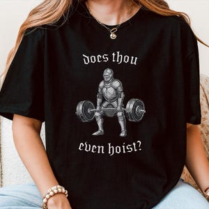 Pode incluir: Camiseta preta com um gráfico de um cavaleiro levantando uma barra. O texto "does thou even hoist?" é impresso acima e abaixo da ilustração do cavaleiro em branco, em uma fonte de estilo gótico. O design é centralizado na camiseta.