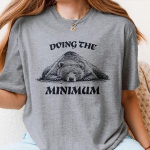 Puede incluir: Camiseta gris con la frase "DOING THE MINIMUM" sobre un gráfico de un oso durmiendo. La camiseta es de cuello redondo y manga corta. El texto y el gráfico del oso son negros.