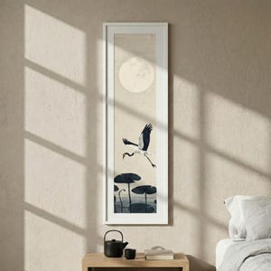 May include: A framed vertical print featuring a crane in flight over a pond with lily pads, under a full moon. The artwork is in shades of beige, white, and black, with a minimalist aesthetic. The frame is white.