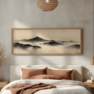 May include: A framed landscape art print depicting a mountain range shrouded in mist. The artwork features a sepia-toned color palette with dark mountain silhouettes against a hazy background. The frame is made of light-colored wood.