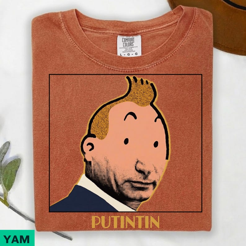 May include: A rust-coloured short-sleeve t-shirt featuring a graphic print of a cartoon character with a prominent quiff hairstyle, wearing a dark blue suit jacket. Below the graphic, the text PUTINTIN is printed in bold, yellow capital letters.