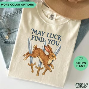 May include: An ivory t-shirt featuring a graphic of a brown rabbit leaping in front of three silver swords. The text "MAY LUCK FIND YOU" is printed in dark blue above the rabbit. The shirt has a crew neckline and a Comfort Colors brand label.