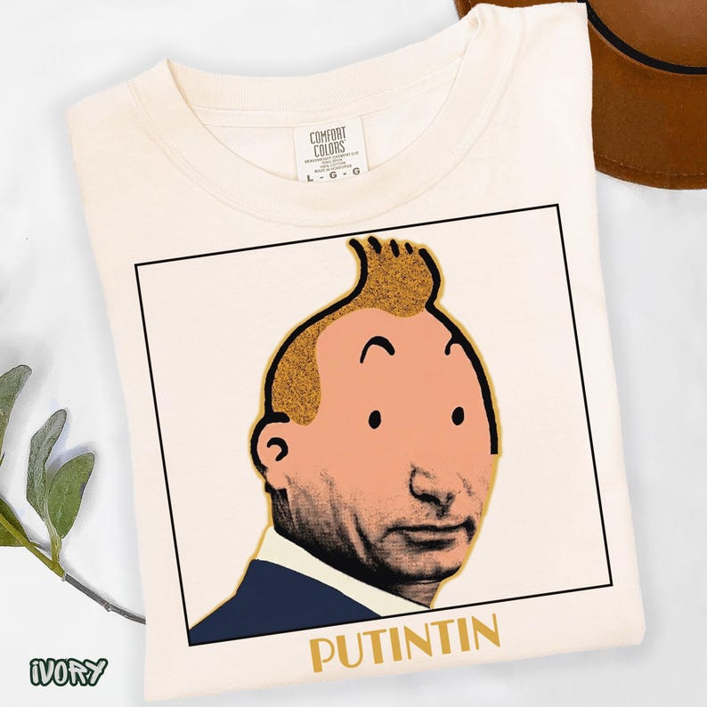 May include: An ivory-coloured t-shirt featuring a square graphic print of a cartoon-style character with a distinctive quiff hairstyle, blending a stylised face with a realistic photographic chin and mouth, with the text PUTINTIN printed below.