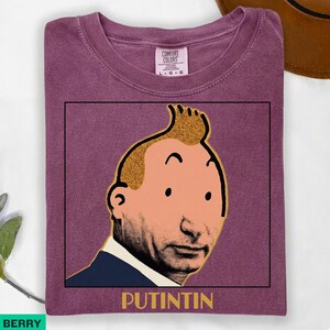 May include: A berry-coloured t-shirt featuring a graphic print of a cartoon-style head with a prominent quiff hairstyle, placed above the text "PUTINTIN" in a bold, yellow, sans-serif font. The shirt has a crew neckline and a Comfort Colors label.