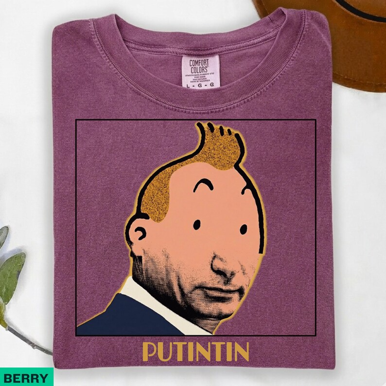May include: A berry-coloured t-shirt featuring a graphic print of a cartoon-style head with a prominent quiff hairstyle, placed above the text "PUTINTIN" in a bold, yellow, sans-serif font. The shirt has a crew neckline and a Comfort Colors label.