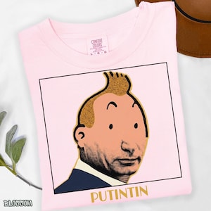 May include: A light pink t-shirt featuring a graphic print of a cartoon-style head with a signature quiff hairstyle, set within a black square frame. Below the illustration, the text PUTINTIN is printed in gold capital letters.
