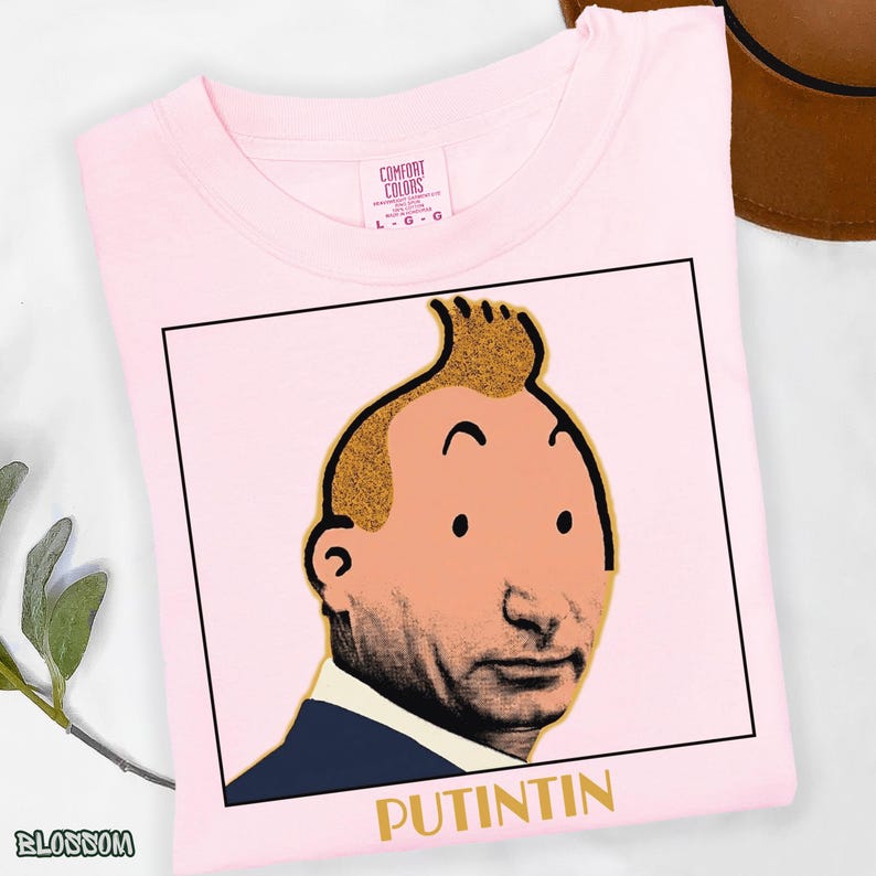 May include: A light pink t-shirt featuring a graphic print of a cartoon-style head with a signature quiff hairstyle, set within a black square frame. Below the illustration, the text PUTINTIN is printed in gold capital letters.