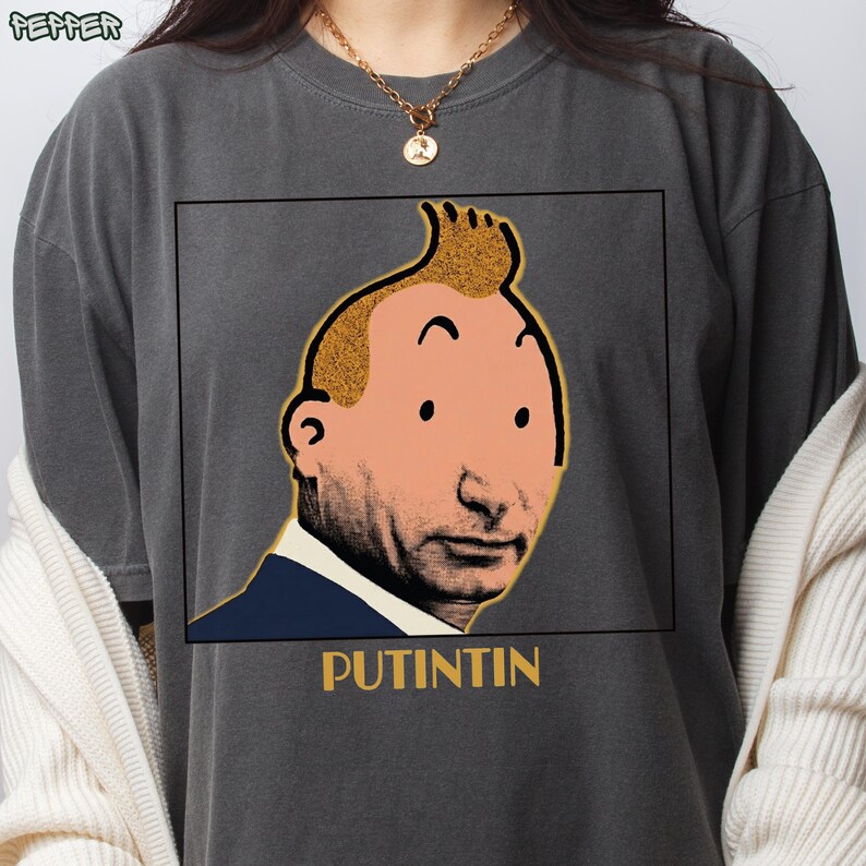 May include: A charcoal grey t-shirt featuring a graphic print of a face with a cartoon-style quiff hairstyle, set within a black square frame. Below the frame, the text PUTINTIN is printed in yellow. The shirt has a crew neckline and short sleeves.