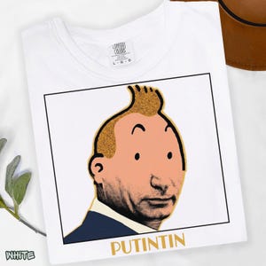May include: A white t-shirt featuring a graphic print of a face with a prominent quiff hairstyle, combining features of a cartoon character and a real person. The design is enclosed in a black square frame with the text PUTINTIN printed in gold below.