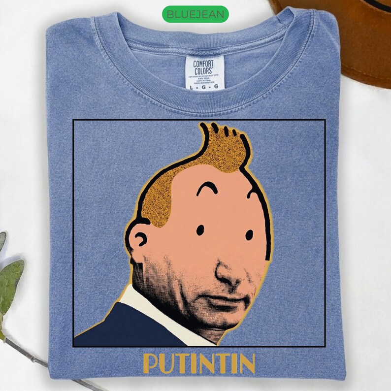 May include: A blue crew neck t-shirt featuring a graphic print of a cartoon character with a distinctive quiff hairstyle combined with a realistic facial portrait. The text PUTINTIN is printed in yellow below the square graphic frame.