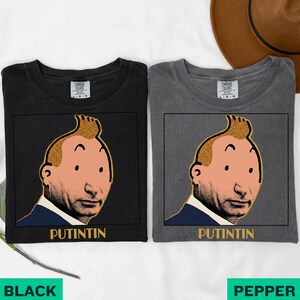 May include: Two short-sleeve crew neck t-shirts, one black and one charcoal grey, each featuring a graphic print of a cartoon-style face with a quiff hairstyle and the text PUTINTIN printed in yellow capital letters below the illustration.