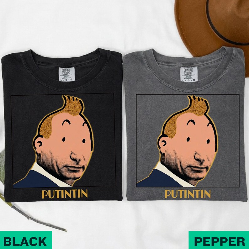 May include: Two short-sleeve crew neck t-shirts, one black and one charcoal grey, each featuring a graphic print of a cartoon-style face with a quiff hairstyle and the text PUTINTIN printed in yellow capital letters below the illustration.