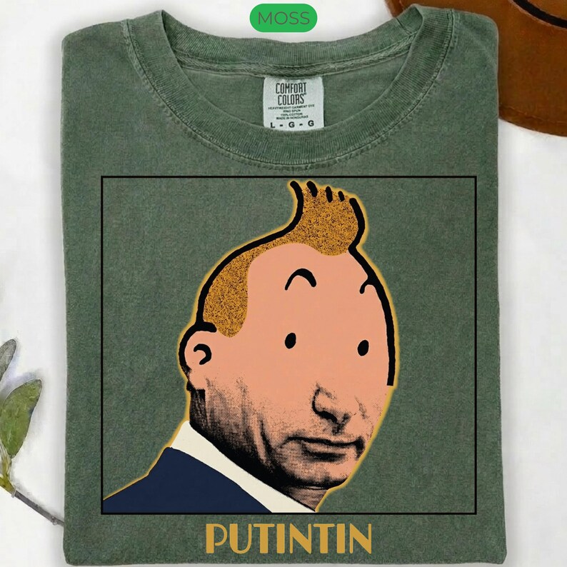 May include: A moss green t-shirt featuring a graphic print of a cartoon character with a distinctive quiff hairstyle and a realistic face. The design is enclosed in a black square frame with the text PUTINTIN printed in yellow below the image.