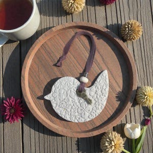 May include: A white ceramic bird ornament with a floral pattern, a purple ribbon, and a small key charm. The ornament rests on a wooden tray with a cup of tea and decorative flowers.